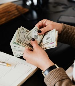 Unrecognizable elegant female in sweater counting dollar bills while sitting at wooden table with planner and pen
